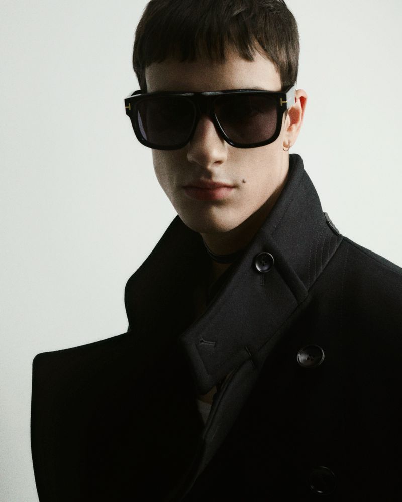 Portrait of a young adult man wearing sunglasses and a dark pea coat against a white background in a studio with professional lighting.