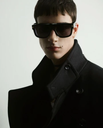 Portrait of a young adult man wearing sunglasses and a dark pea coat against a white background in a studio with professional lighting.