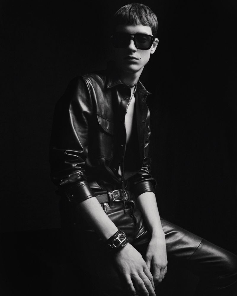 Monochrome portrait of a young adult man in a leather shirt and pants, seated in a dark studio wearing sunglasses and a bracelet.