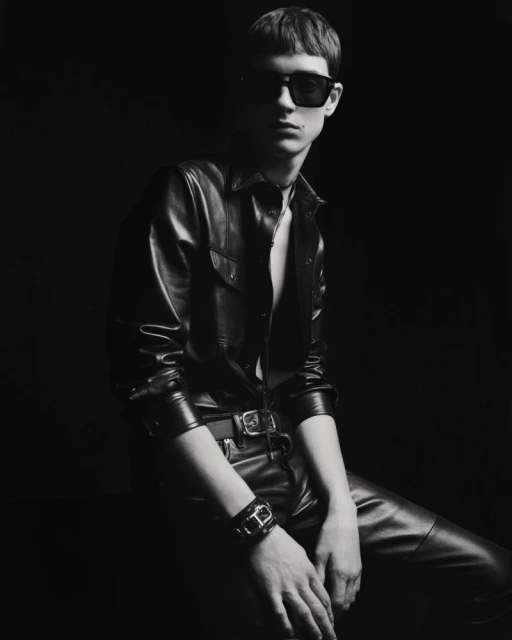 Monochrome portrait of a young adult man in a leather shirt and pants, seated in a dark studio wearing sunglasses and a bracelet.
