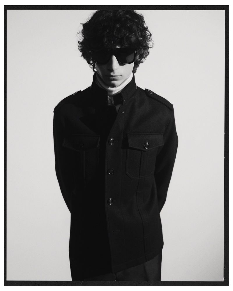 A young adult male with curly hair and sunglasses poses indoors wearing a black jacket and white turtleneck against a white background.