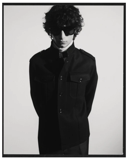 A young adult male with curly hair and sunglasses poses indoors wearing a black jacket and white turtleneck against a white background.