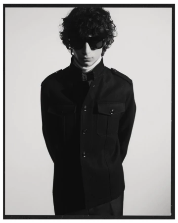 A young adult male with curly hair and sunglasses poses indoors wearing a black jacket and white turtleneck against a white background.
