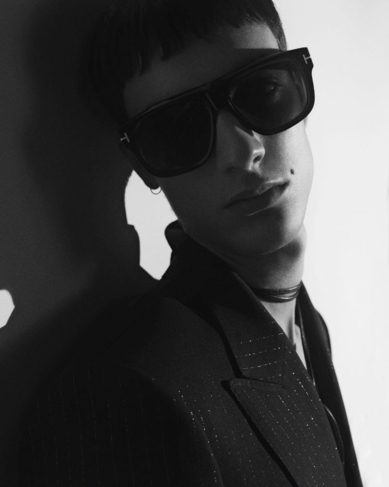 Close-up portrait of a male model wearing dark oversized sunglasses and a black blazer against a light wall.