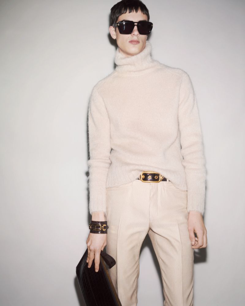 A light-skinned male model wears a beige turtleneck, pants, sunglasses and a brown leather clutch against a plain light grey background.