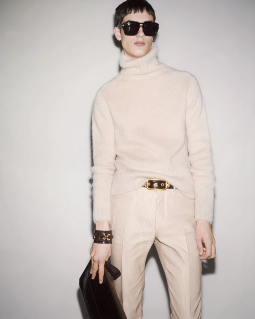 A light-skinned male model wears a beige turtleneck, pants, sunglasses and a brown leather clutch against a plain light grey background.