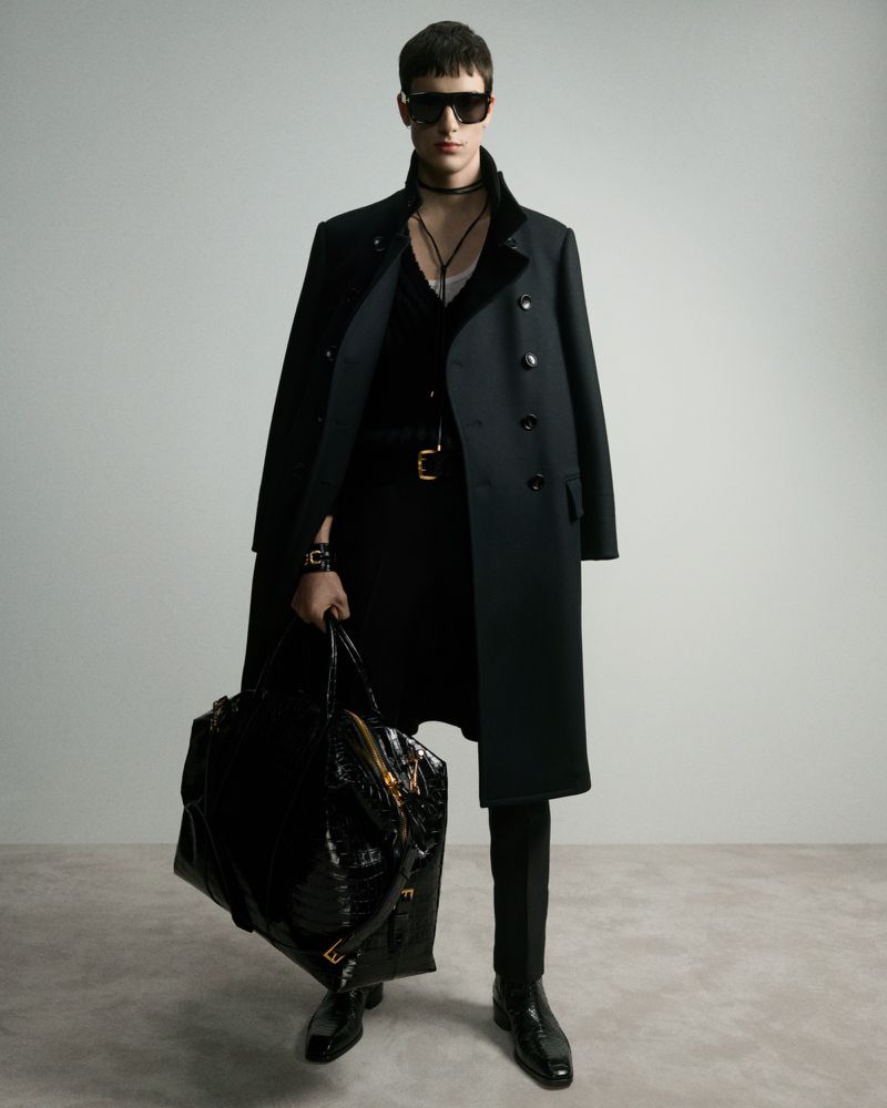 A male model in a black coat and sunglasses poses in a fashion shot, holding a large textured bag against a neutral backdrop.