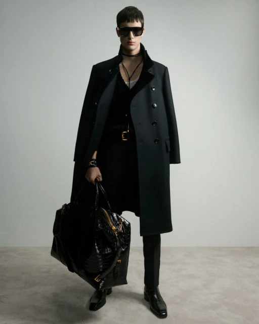 A male model in a black coat and sunglasses poses in a fashion shot, holding a large textured bag against a neutral backdrop.