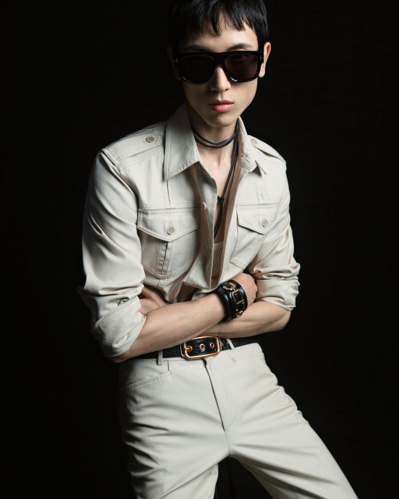 Person in a beige outfit and black sunglasses posing with arms crossed against a dark background in a fashion forward portrait.