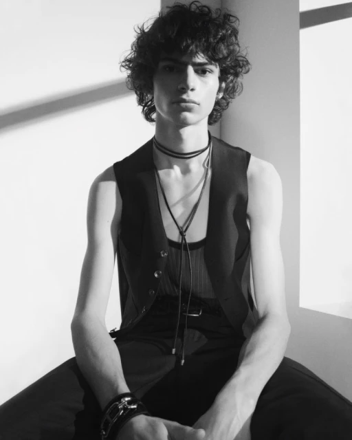 Monochrome portrait of a curly-haired male model in a dark vest and pants, seated against a bright wall with a shadow.
