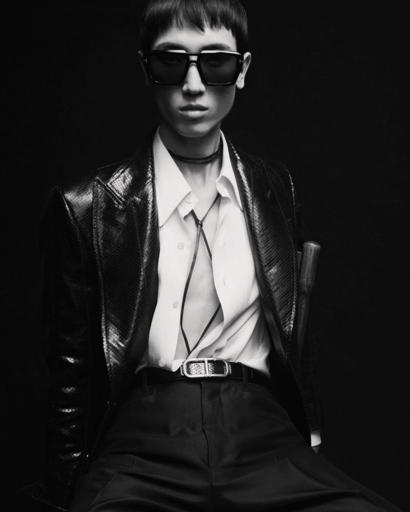 A person poses in black pants, snakeskin jacket, white shirt, and sunglasses against a dark background for a fashion editorial.