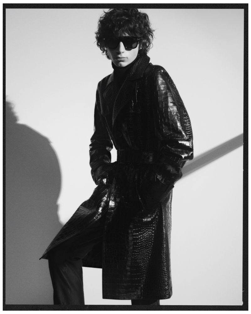 Monochrome fashion shot of a male model with curly hair wearing sunglasses and an embossed crocodile-pattern coat, posing in a photo studio.