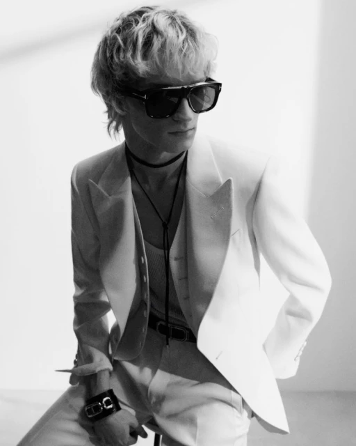 Black and white studio portrait of a fair-skinned adult male wearing a white suit, tank top, bolo tie, sunglasses, and wrist cuff.