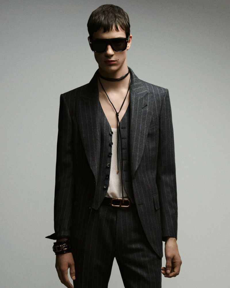 Fashionable young adult man in a pinstripe suit, vest, and sunglasses posing in a studio with a grey background. He wears jewelry and a belt.