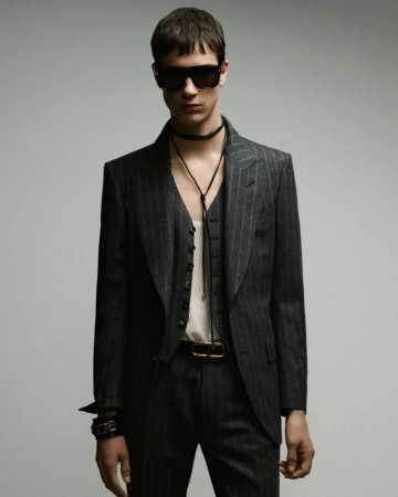 Fashionable young adult man in a pinstripe suit, vest, and sunglasses posing in a studio with a grey background. He wears jewelry and a belt.