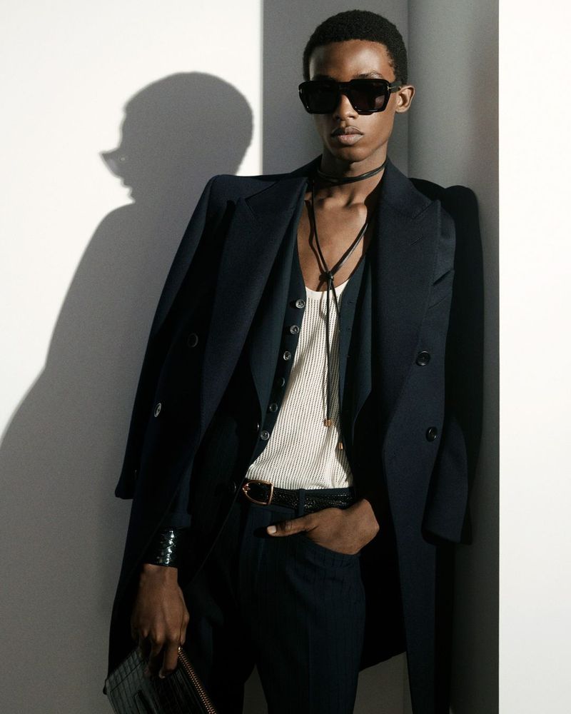 Model in dark sunglasses wearing a black overcoat and white ribbed vest leaning against a wall