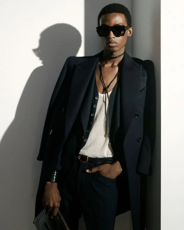 Model in dark sunglasses wearing a black overcoat and white ribbed vest leaning against a wall