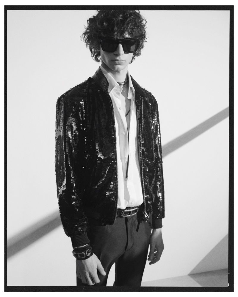 A male model wearing a black sequin bomber jacket and dark sunglasses against a plain white studio backdrop