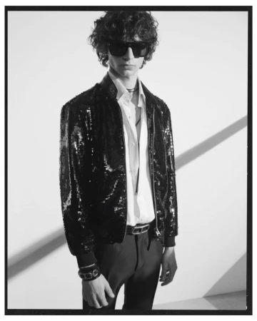 A male model wearing a black sequin bomber jacket and dark sunglasses against a plain white studio backdrop