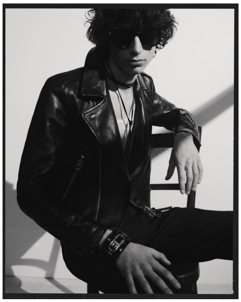 Monochrome portrait of a young adult with curly hair, wearing a leather jacket, sunglasses, sitting in a chair against a light background.