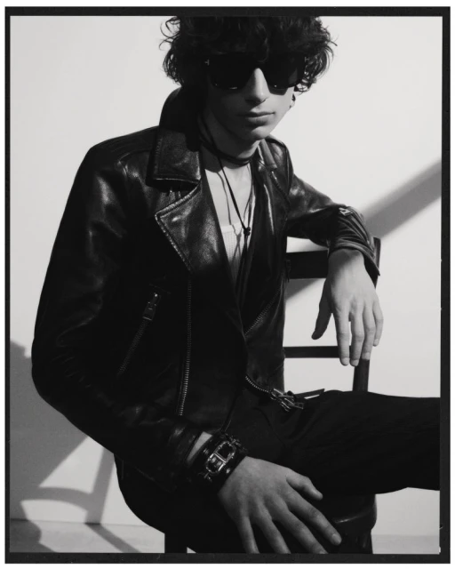 Monochrome portrait of a young adult with curly hair, wearing a leather jacket, sunglasses, sitting in a chair against a light background.