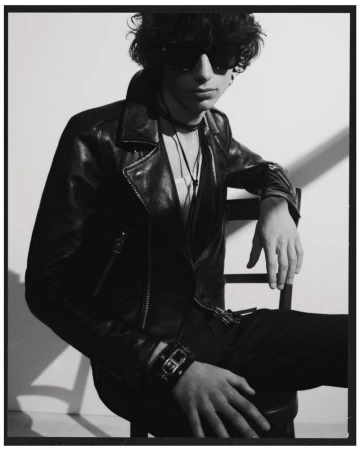 Monochrome portrait of a young adult with curly hair, wearing a leather jacket, sunglasses, sitting in a chair against a light background.
