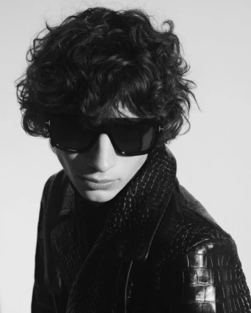Close-up portrait of a male model wearing dark sunglasses and a black textured leather jacket against a light background.