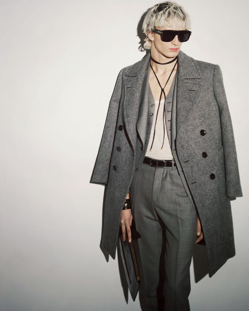 Stylish adult male model posing indoors wearing a grey wool coat, vest, trousers, and sunglasses against a white backdrop.