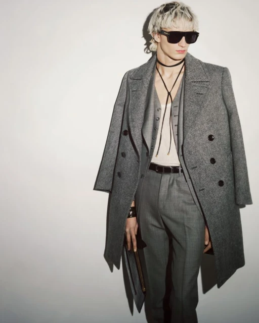 Stylish adult male model posing indoors wearing a grey wool coat, vest, trousers, and sunglasses against a white backdrop.