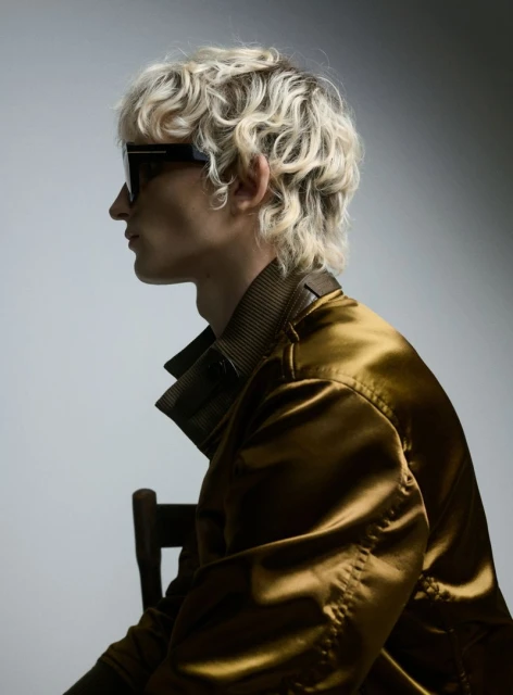 A person with wavy blond hair sits wearing sunglasses and a gold jacket against a light grey background, with a portion of a wooden chair visible.