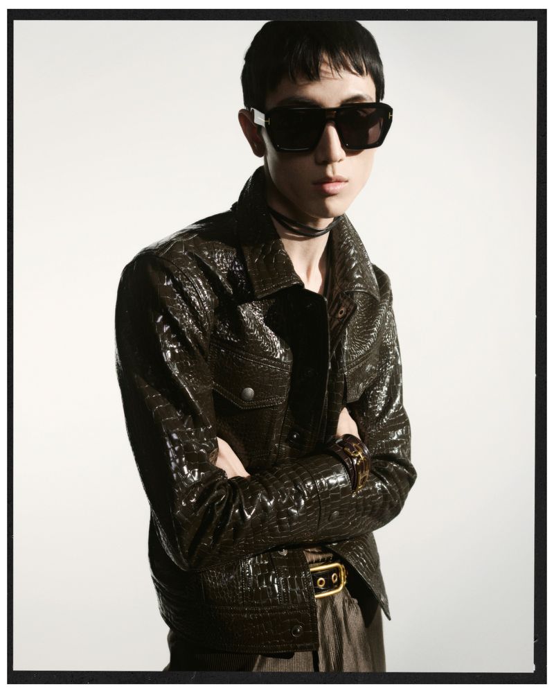 Portrait of a young adult male with dark hair wearing sunglasses and a crocodile-textured jacket in front of a bright background.