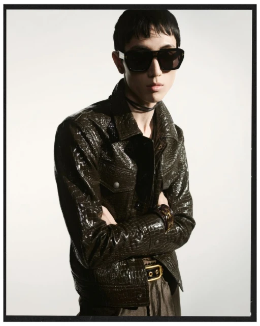 Portrait of a young adult male with dark hair wearing sunglasses and a crocodile-textured jacket in front of a bright background.