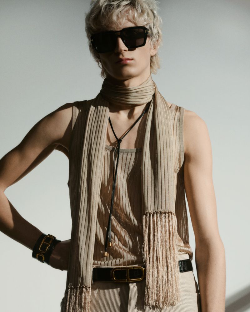 Man poses in sunglasses, wearing a ribbed tank, scarf, and belt. The scene is brightly lit against a neutral background.