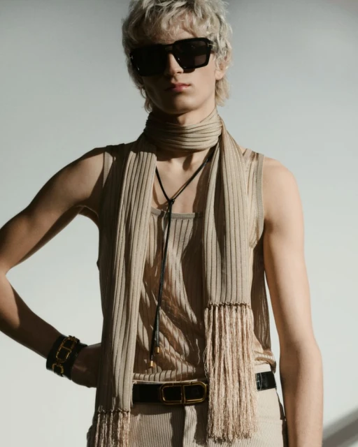 Man poses in sunglasses, wearing a ribbed tank, scarf, and belt. The scene is brightly lit against a neutral background.