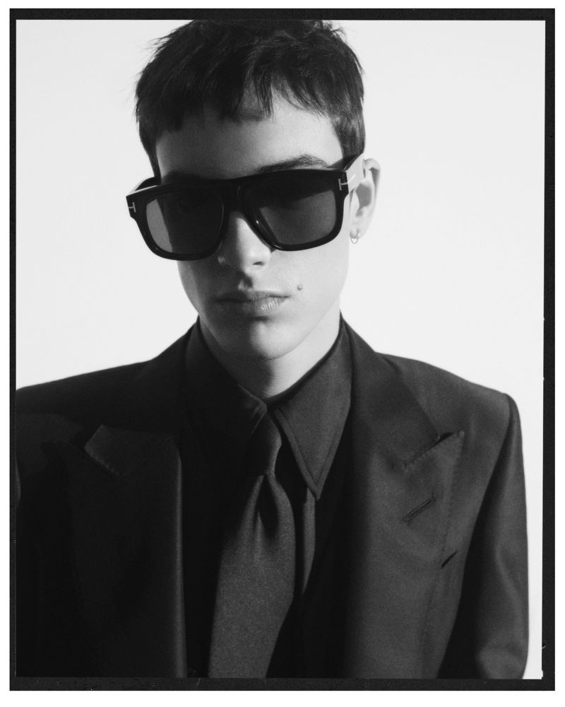 Portrait of a young male model wearing black sunglasses, a tailored dark suit jacket, and matching tie against a clean background