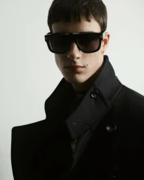 Close-up portrait of a male model wearing dark oversized sunglasses and a black collared coat against a plain white background.