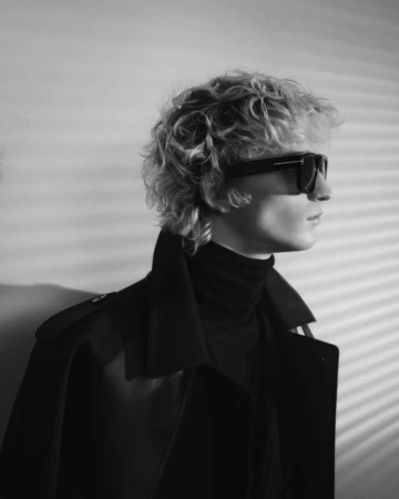 Profile portrait of a male model wearing dark sunglasses and a black coat against striped slatted light shadows.