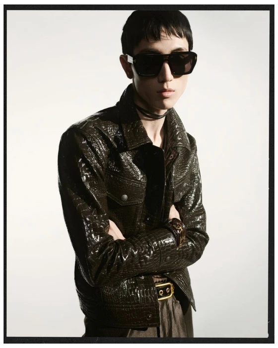 Model wearing dark sunglasses and a textured brown leather jacket against a seamless white background.