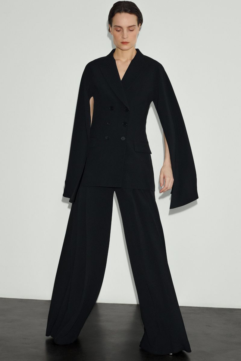 A woman in a black pantsuit with cape-like sleeves stands against a white background in a fashion shot, showcasing a modern design.