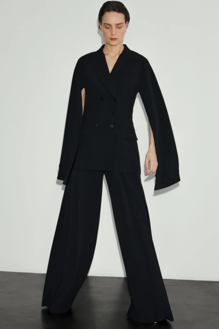 A woman in a black pantsuit with cape-like sleeves stands against a white background in a fashion shot, showcasing a modern design.