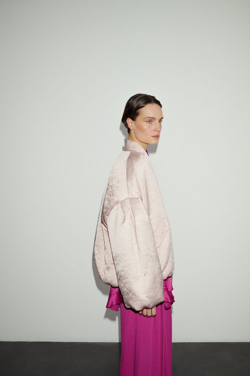 Woman in blush jacket and fuchsia pants poses in a studio. Minimalist background highlights fashion and textures in soft, natural light.