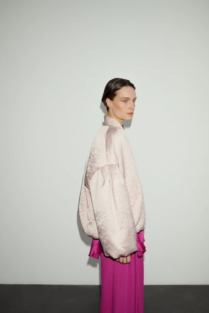 Woman in blush jacket and fuchsia pants poses in a studio. Minimalist background highlights fashion and textures in soft, natural light.