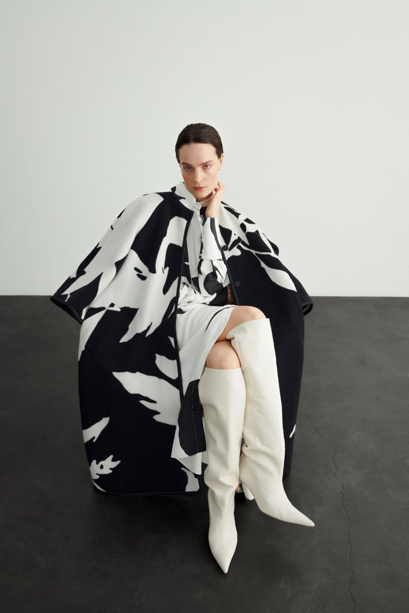 Fashion photo of a woman sitting and posing in a black and white patterned coat and white boots in front of a grey backdrop.
