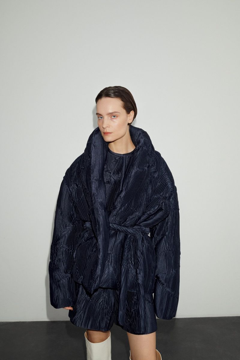 Woman wearing a navy blue, quilted, oversized jacket with a belt and matching shorts, posed in front of a solid white background.