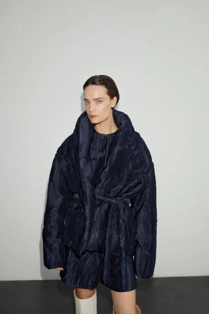 Woman wearing a navy blue, quilted, oversized jacket with a belt and matching shorts, posed in front of a solid white background.