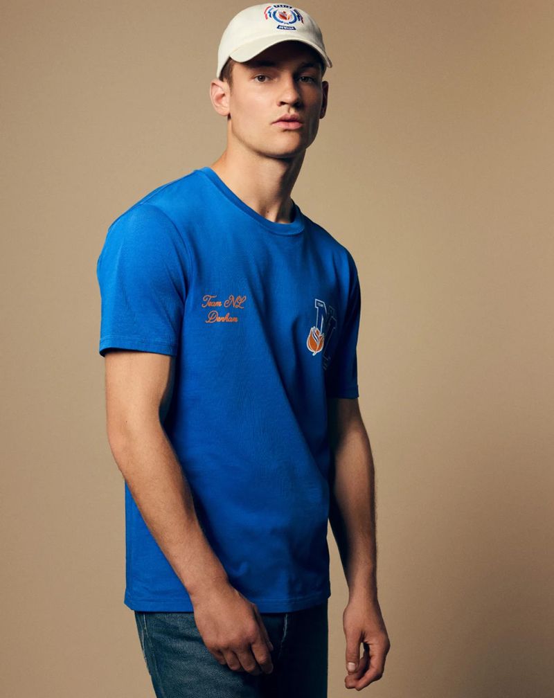 A young adult man in a blue t-shirt and cap poses in front of a neutral background, showcasing a casual, sporty fashion style.