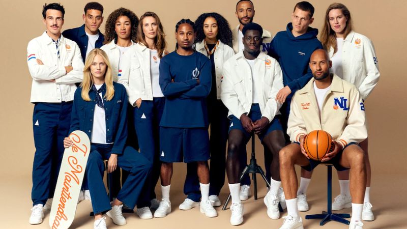 Group of diverse adults and young adults posing in matching blue and white athletic wear indoors, some holding a skateboard and basketball.