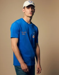 Model wearing a blue t-shirt, blue jeans, and a white baseball cap against a solid warm beige studio background.