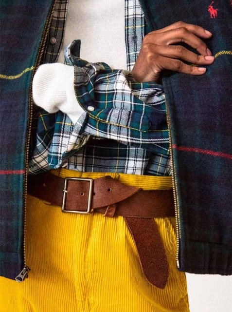 Close-up of a light-skinned person wearing a plaid jacket, plaid shirt, white sweater, belt, and yellow pants in an indoor setting.