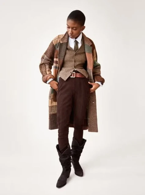 A woman in a brown tweed suit and coat poses in front of a neutral background. She is wearing black boots and a brown belt.